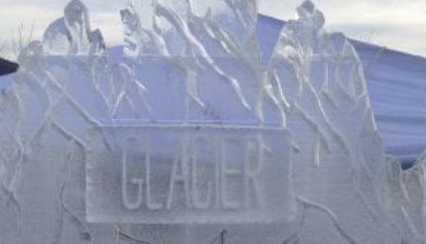 Ice sculpture with the word GLACIER carved in its center. The sculpture features figures of people running above the text. A tent and the sky are visible in the background.