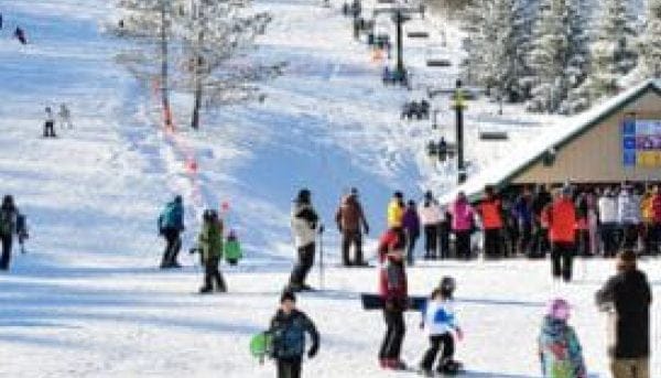 A snowy ski resort scene with children and adults skiing and snowboarding. People gather near a lodge, with snow-covered trees in the background. The sky is clear, and the atmosphere is lively and active.