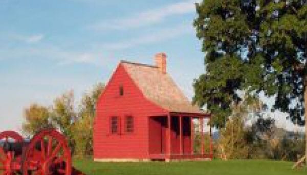 A small red wooden house with a chimney sits on a grassy field beneath the blue skies of Saratoga Springs. To the right, a large green tree provides shade, while an old-fashioned red cartwheel rests quietly to the left.