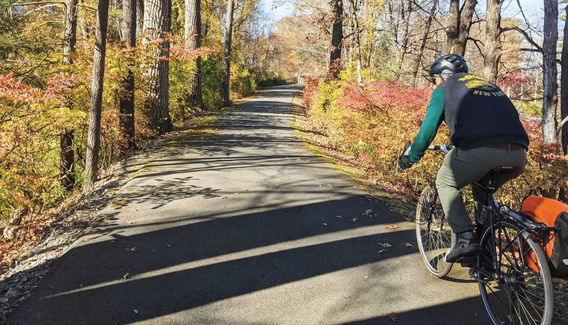 A person rides a bicycle along the Empire State Trail, surrounded by autumn trees with colorful leaves. The sun casts long shadows across the paved path. The rider, wearing a helmet and a jacket, has an orange bag attached to the bike.
