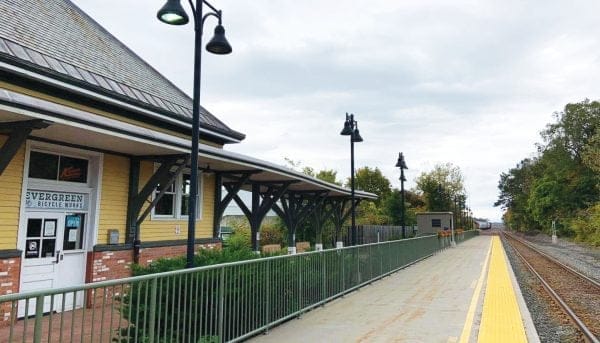A quaint train station platform with a yellow and brick building on the left evokes memories of the Empire State Trail. Street lamps line the platform, and railroad tracks stretch into the distance. Trees and cloudy skies frame this picturesque setting.