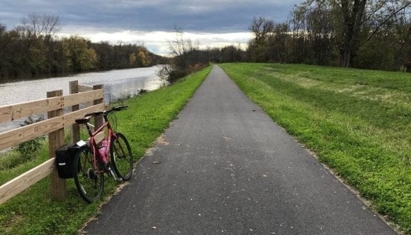The Empire State Trail features a paved bike path meandering along a calm river, bordered by wooden fencing and lush grassy areas. A bike rests against the fence on the left, while trees line the path under a cloudy sky.