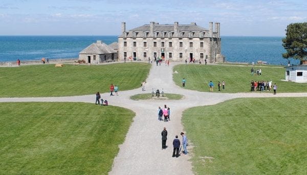A historic stone fort with a symmetrical design overlooks the blue expanse under a clear sky, reminiscent of the power and grandeur of Niagara Falls. Green lawns with crossing paths lead to the entrance, where people are scattered across the grounds, savoring the scenic beauty.
