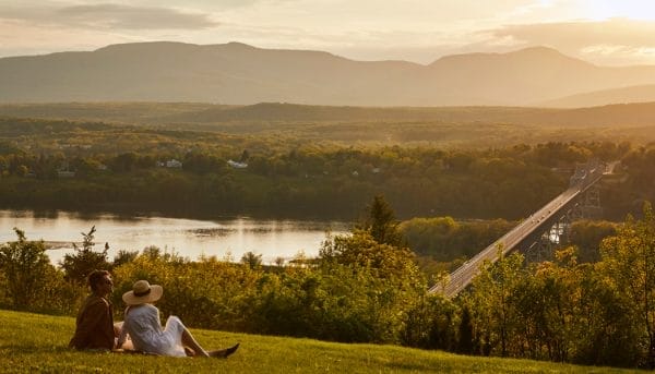 A couple sits on a grassy hill in the Hudson Valley, gazing at a scenic view during sunset. A river flows below, with a long bridge crossing it. The distant landscape is filled with trees and silhouetted mountains under a golden sky.