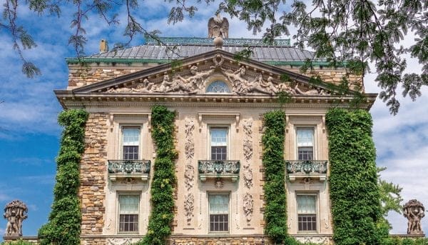 A grand historic building in Hudson Valley boasts a stone facade, intricate sculptures, and ornate carvings. Ivy partially covers the walls. The structure features large windows and a decorative triangular pediment, all framed by a bright blue sky and leafy branches.