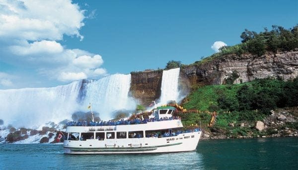 A tour boat, filled with passengers, navigates the waters near a large, cascading waterfall under a clear blue sky. Lush greenery surrounds the rocky cliffs of Niagara Falls, and the boats name is visible on its side.
