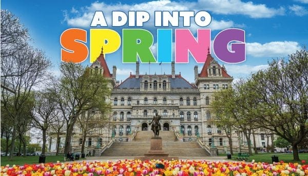 An ornate building with a staircase, surrounded by trees and colorful tulips in the foreground, evokes the charm of the Empire State Trail. The sky is blue with clouds. Large text at the top reads A Dip Into Spring in vibrant colors.