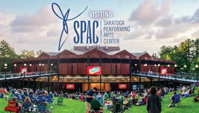 On a dusky evening, a crowd gathers on the lawn before the illuminated Saratoga Performing Arts Center. The iconic SPAC logo shines above, alongside the welcoming text: Visiting Saratoga Performing Arts Center.