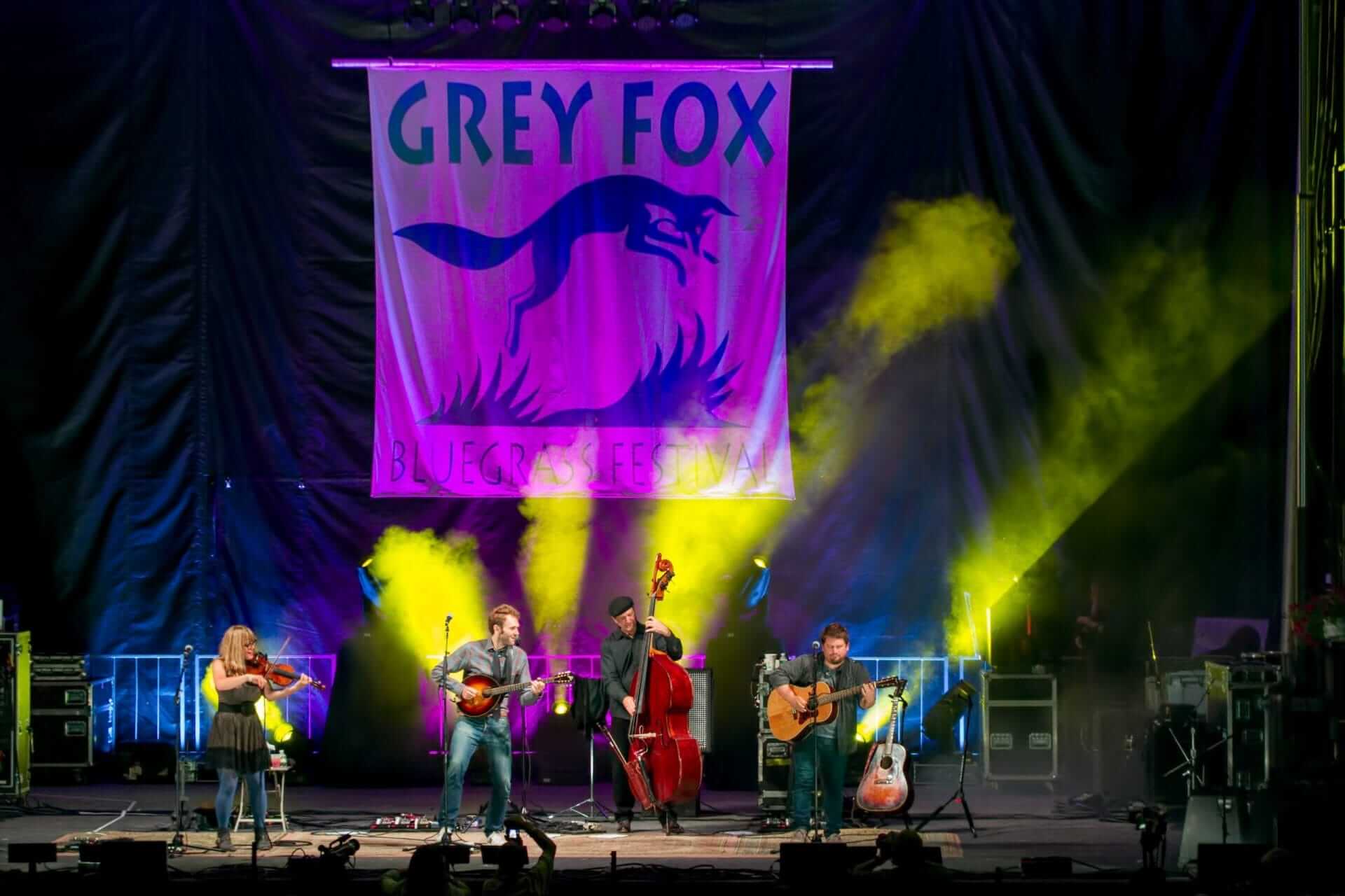 The Grey Fox Bluegrass Festival comes alive as a band performs on stage. Four musicians skillfully play the fiddle, guitar, mandolin, and upright bass while vibrant yellow and green lights illuminate the energetic scene.