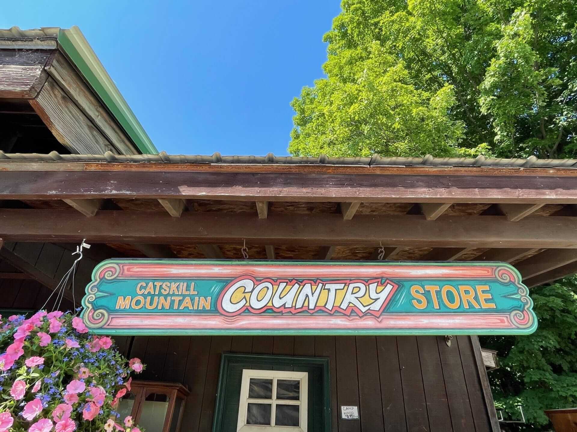 A wooden sign for Catskill Mountain Country Store hangs under a rustic roof. Pink flowers in a basket appear to the left, as if welcoming visitors to the nearby Grey Fox Bluegrass Festival. A green tree and bright blue sky complete the charming backdrop.