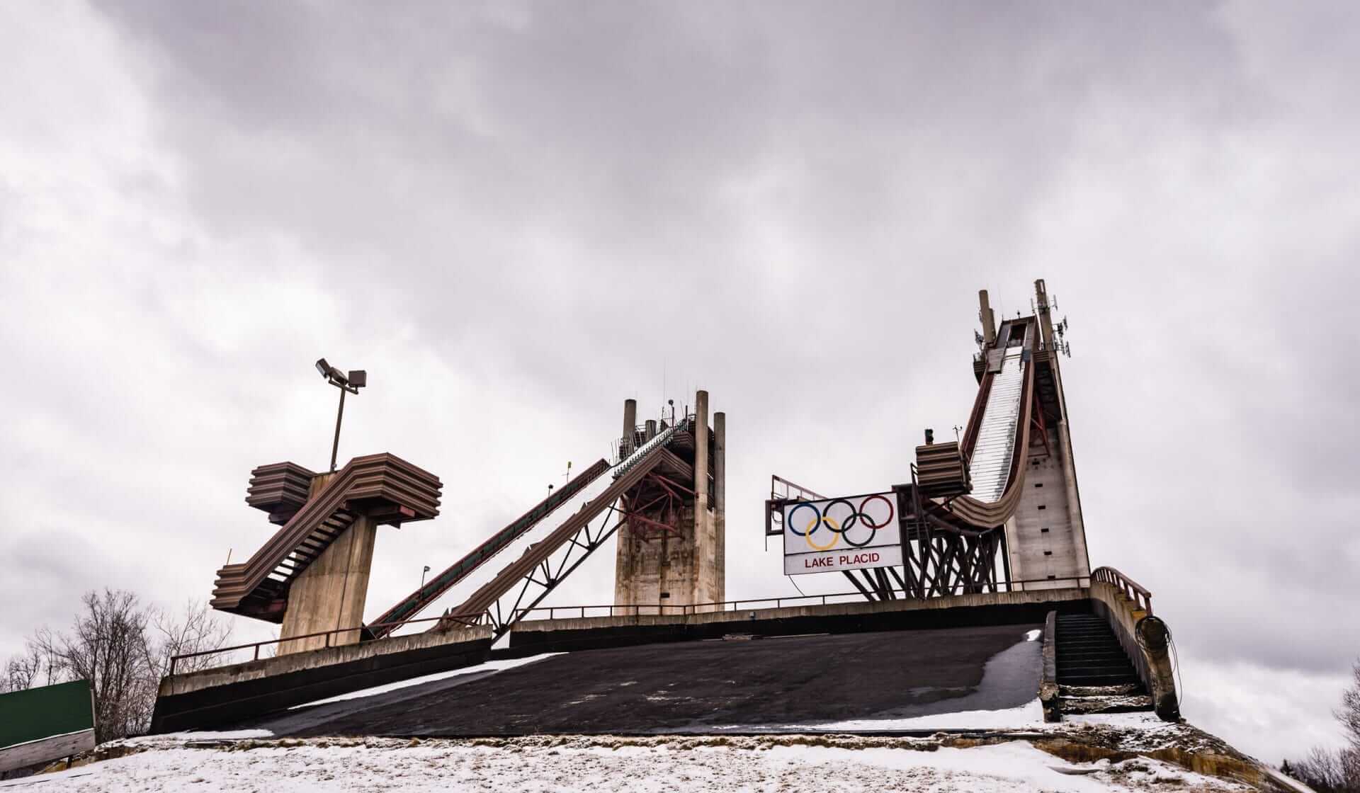 Under a cloudy sky, the Lake Placid ski jump complex stands tall, its ramps with red supports resembling majestic giants. Stairs ascend to the top, echoing the ambition of athletes from places like Yonkers. The Olympic rings and Lake Placid sign beam proudly as snow blankets the ground.