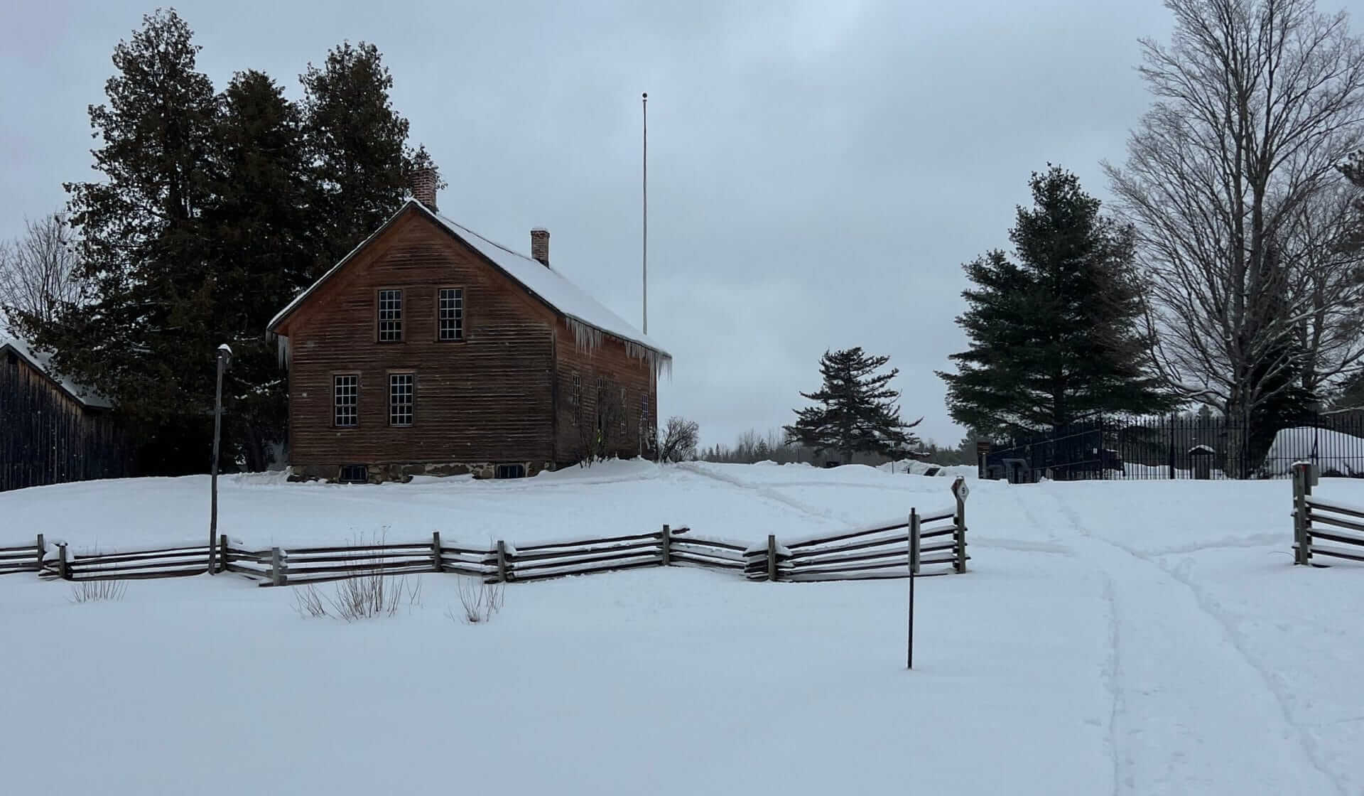 A wooden house stands in a snowy Yonkers landscape, surrounded by trees and a wooden fence. The sky is overcast, suggesting a cold winter day. A path is visible in the snow leading towards the house.