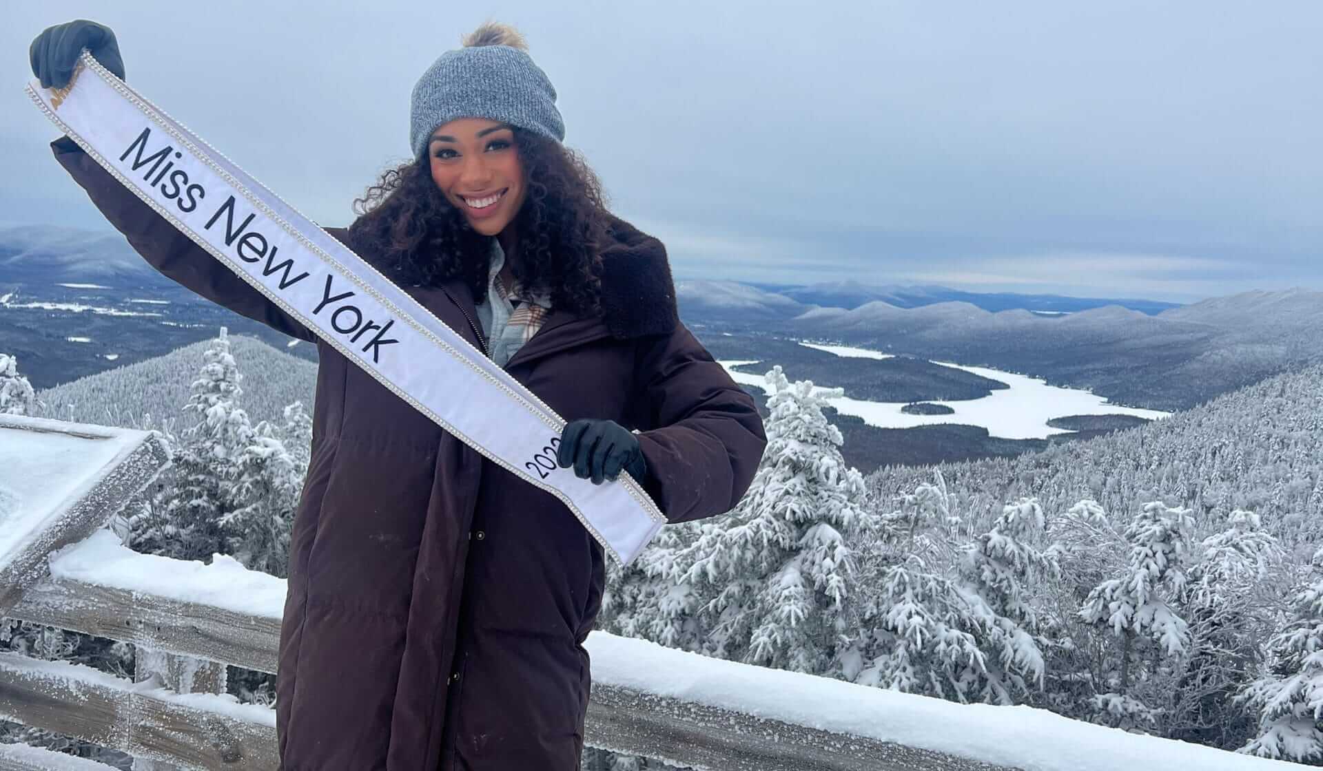 A woman in a winter coat and beanie stands on a snowy mountaintop, smiling and holding a Miss New York sash. Snow-covered trees and the stunning mountain range stretch beyond, capturing a moment that echoes the charm of her hometown, Yonkers.