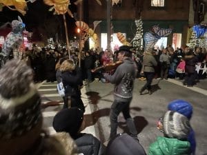 A street performance in Rhinebeck at night with a crowd gathered around. A musician plays the violin while others hold large, colorful animal-shaped lanterns. People wear winter clothing, and buildings with glowing lights are visible in the background.