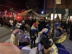 A lively street parade in Rhinebeck at night features people wearing costumes and playing brass instruments. The crowd gathers under festive lights, captivated by a large dragon-shaped puppet leading the joyous procession.