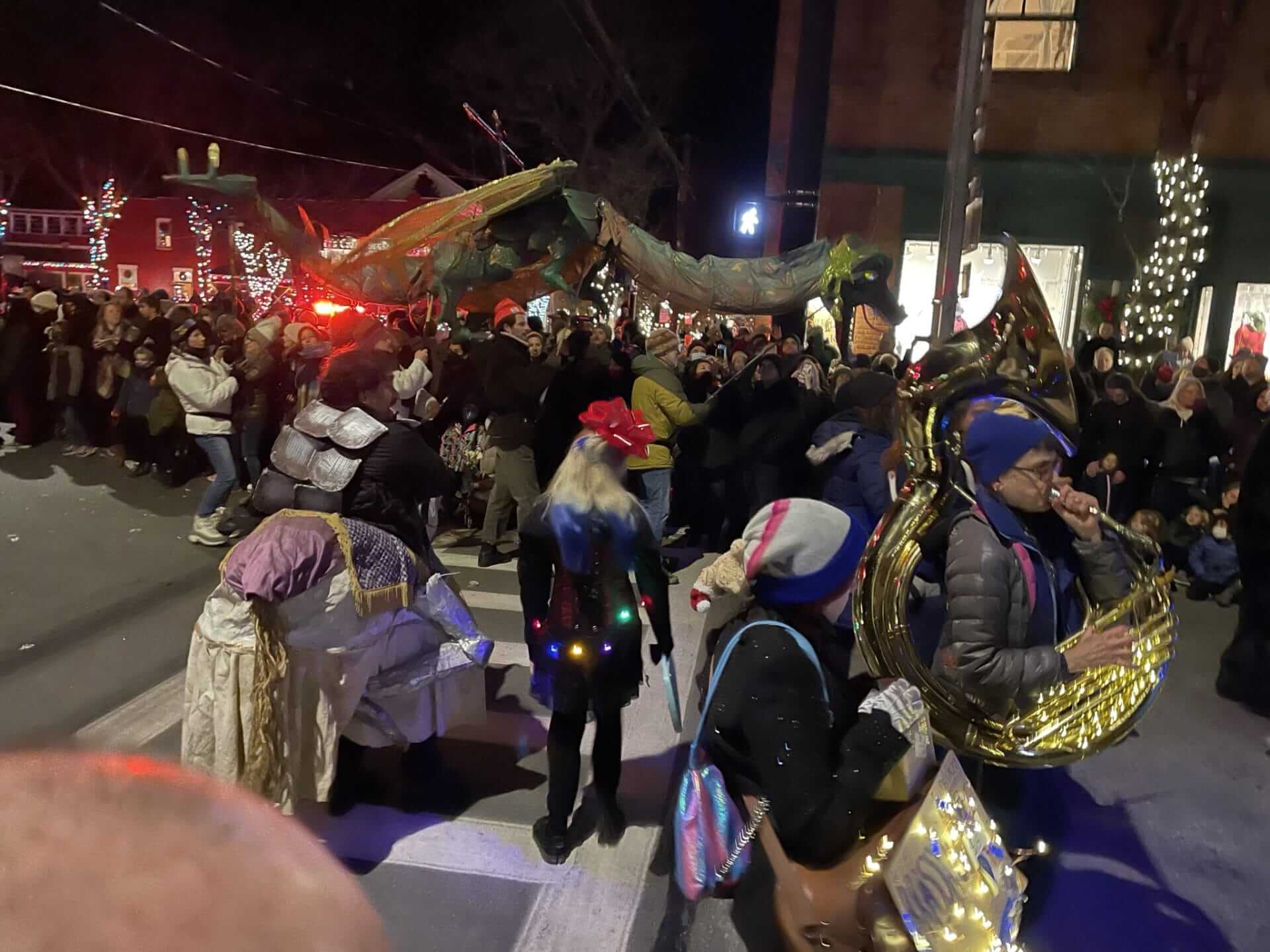 A lively street parade in Rhinebeck at night features people wearing costumes and playing brass instruments. The crowd gathers under festive lights, captivated by a large dragon-shaped puppet leading the joyous procession. A lively street parade in Rhinebeck at night features people wearing costumes and playing brass instruments. The crowd gathers under festive lights, captivated by a large dragon-shaped puppet leading the joyous procession.