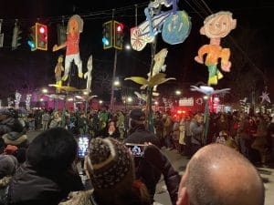 In Rhinebeck, a crowd gathers at night to watch a parade with colorful, oversized puppets resembling children, held high on poles. Bright streetlights and festive lights illuminate the scene, while people eagerly capture the moment with their phones.