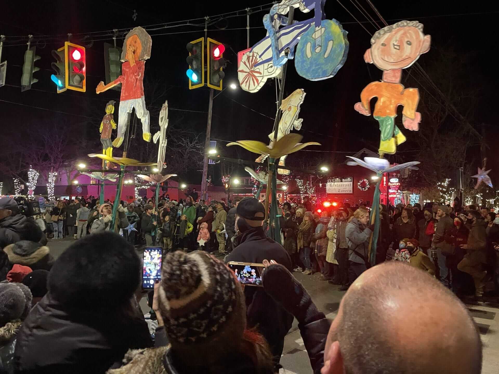 In Rhinebeck, a crowd gathers at night to watch a parade with colorful, oversized puppets resembling children, held high on poles. Bright streetlights and festive lights illuminate the scene, while people eagerly capture the moment with their phones. In Rhinebeck, a crowd gathers at night to watch a parade with colorful, oversized puppets resembling children, held high on poles. Bright streetlights and festive lights illuminate the scene, while people eagerly capture the moment with their phones.