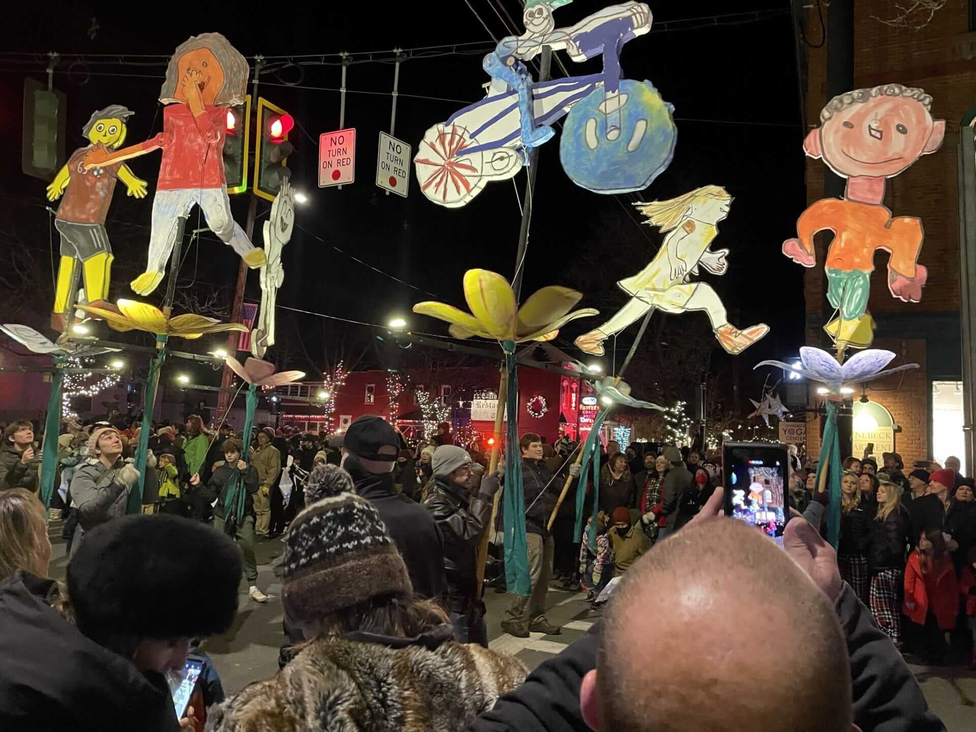 A festive street scene in Rhinebeck unfolds with a crowd gathered around large, colorful, child-like figures held up on poles. The background is adorned with holiday lights and decorations, as some individuals capture the enchanting event on their phones. A festive street scene in Rhinebeck unfolds with a crowd gathered around large, colorful, child-like figures held up on poles. The background is adorned with holiday lights and decorations, as some individuals capture the enchanting event on their phones.