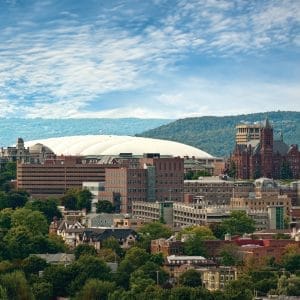 A cityscape featuring a mix of old and modern buildings, lush green trees, and a large, white-domed stadium in the background is perfect for those planning overnight itineraries. The sky is partly cloudy, and a range of hills is visible in the distance.