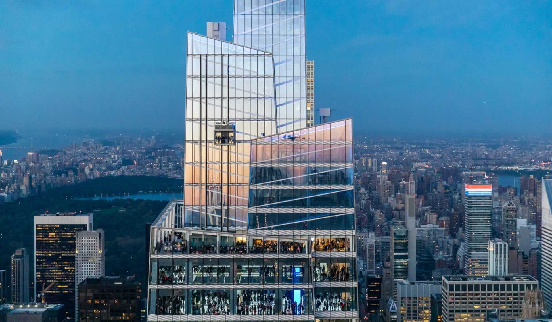 Aerial view of Summit One Vanderbilt, a tall, modern skyscraper with glass facades reflecting the dusky sky. The building reaches new heights with multiple terraces and is surrounded by a sprawling urban landscape, featuring a park visible in the background.