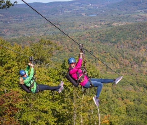 Two people zip-lining down a hill amidst lush green and autumn foliage. They are wearing helmets and harnesses, with expansive forested hills and a mountain range in the background under a clear sky.
