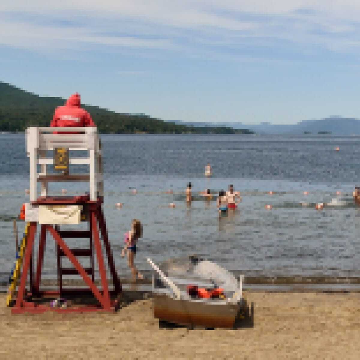 A lifeguard in a red uniform sits on a wooden tower overlooking a beach. Swimmers enjoy the water, with buoys visible in the distance. A small boat rests on the sandy shore. Hills and a cloudy sky form the backdrop.