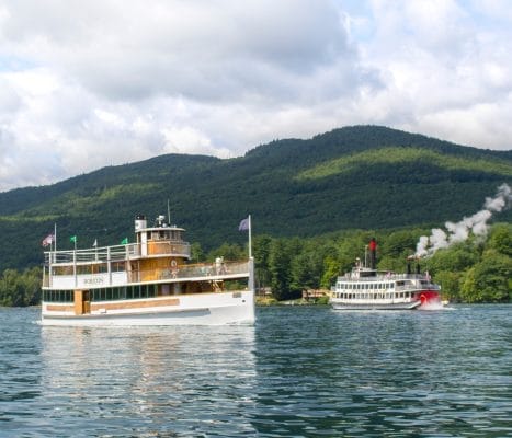 Two vintage steamboats sail on a calm lake surrounded by lush green hills and a partly cloudy sky. One boat is closer, with American flags and a wooden deck, while the other emits steam in the distance.