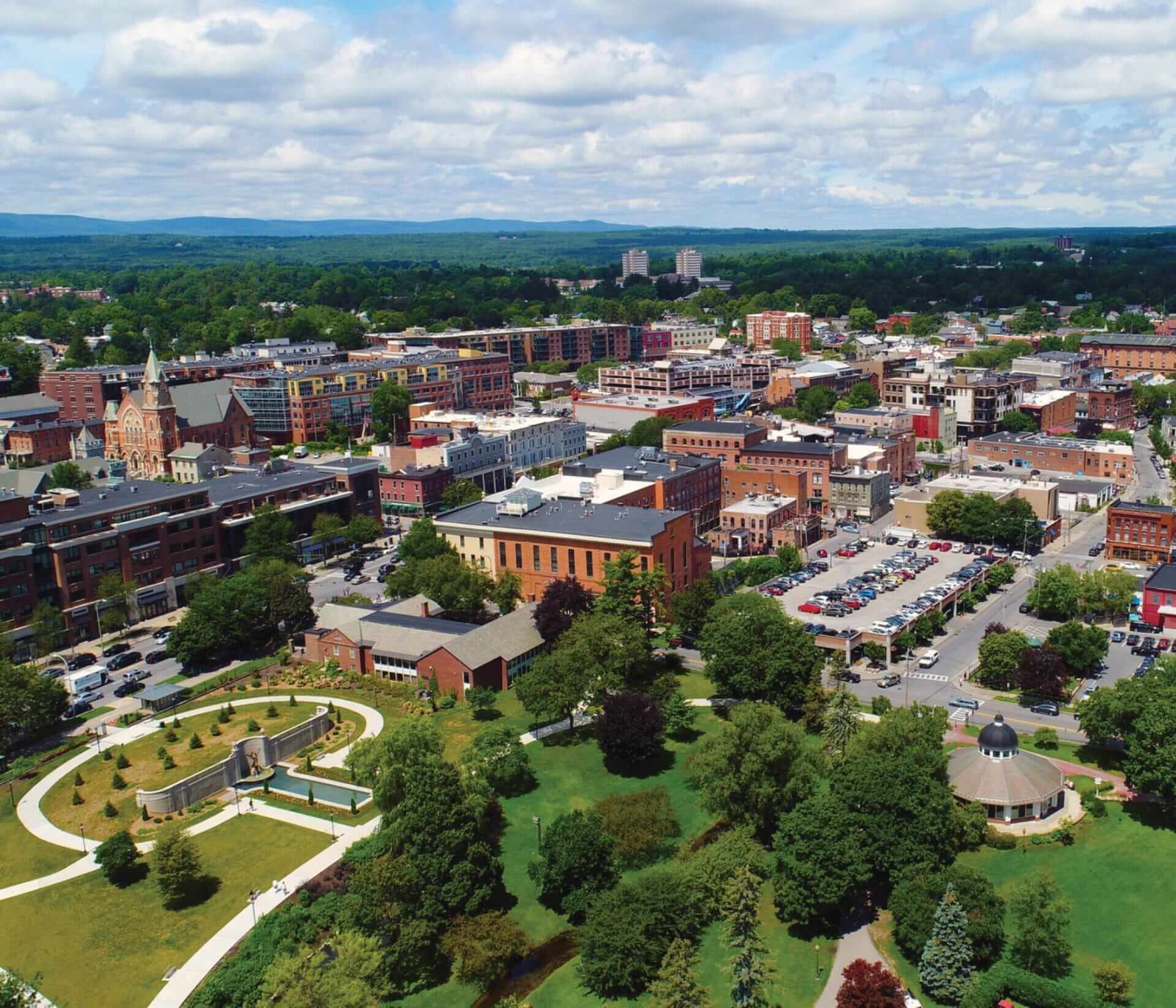 Aerial view of a city with a mix of historic and modern buildings, tree-lined streets, and green spaces. In the foreground, a well-maintained park with pathways and trees is visible. The background shows a hilly landscape under a partly cloudy sky.
