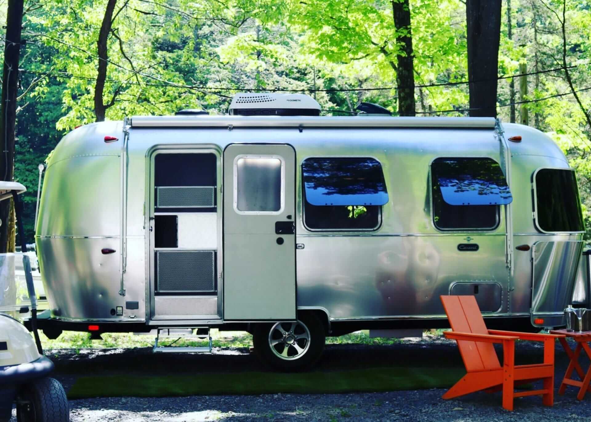 A shiny silver Airstream trailer is parked in a lush, wooded area near Yonkers. Two orange Adirondack chairs sit invitingly on the grass, with part of a white vehicle peeking into view on the left.