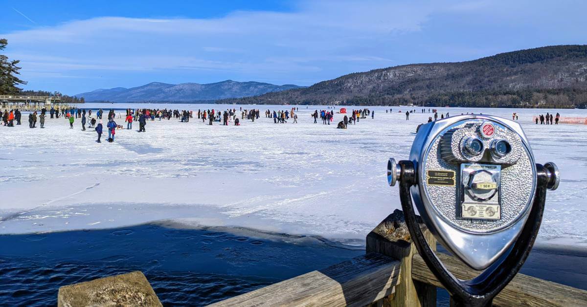 A frozen lake bustling with people enjoying winter events on the ice. In the foreground, a coin-operated viewer invites curious gazes towards snowy mountains and a clear blue sky. Its 2025 in New York State, where trees gracefully outline the shore.
