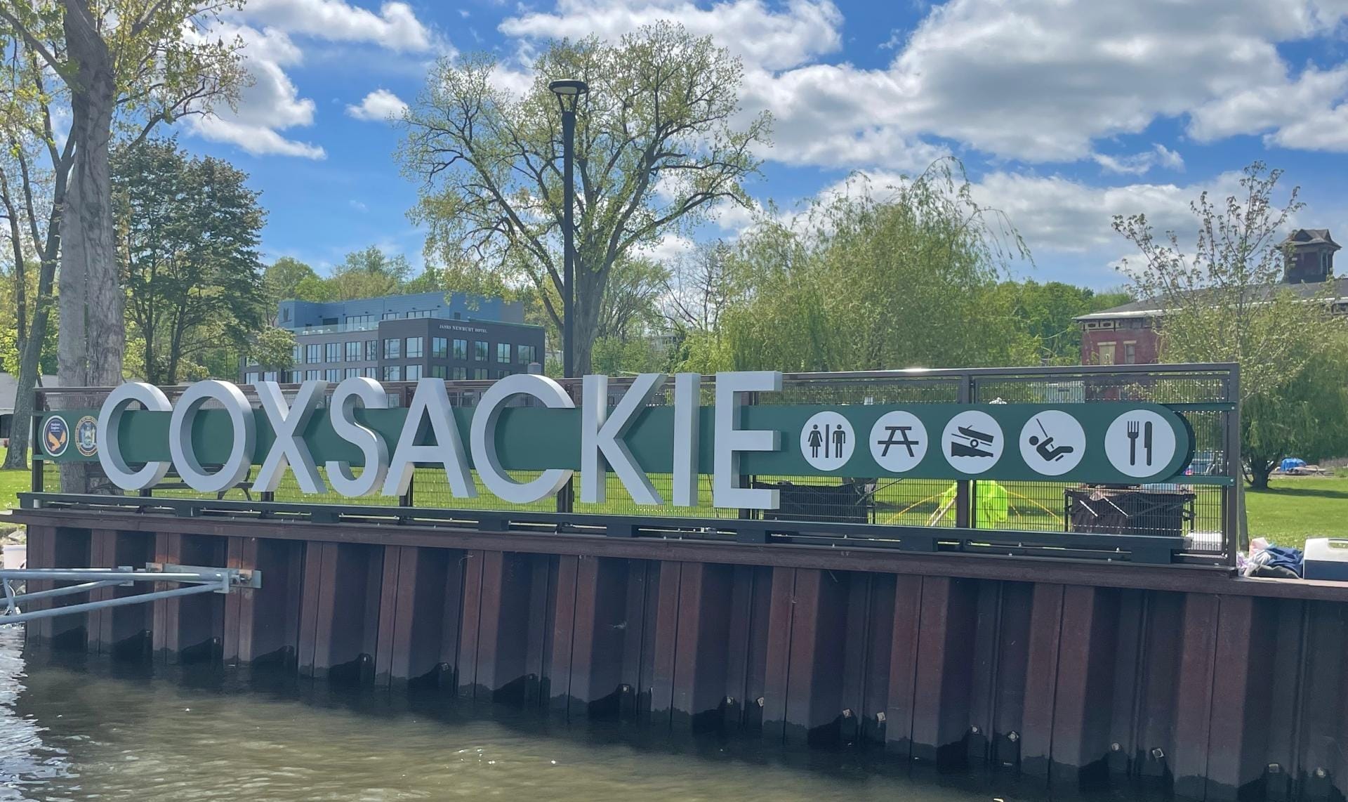 Sign reading Coxsackie on a waterfront pier with icons for amenities such as restrooms, picnic area, and accessibility. A perfect weekend spot in Greene County, framed by trees and buildings under a partly cloudy sky.