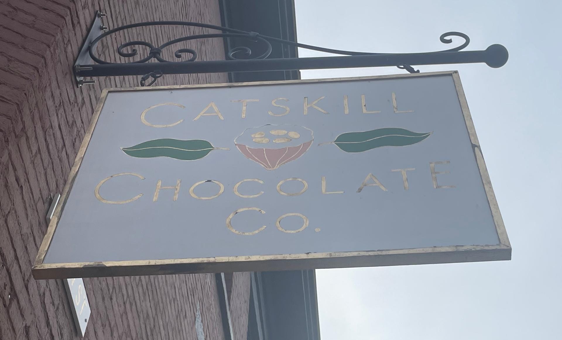 A rectangular sign hanging from a decorative metal bracket on a brick wall. The sign reads Catskill Chocolate Co. in gold letters, with an illustration of a cocoa pod and two leaves in the center—a perfect weekend stop for mastering the art of chocolate enjoyment in Greene County.