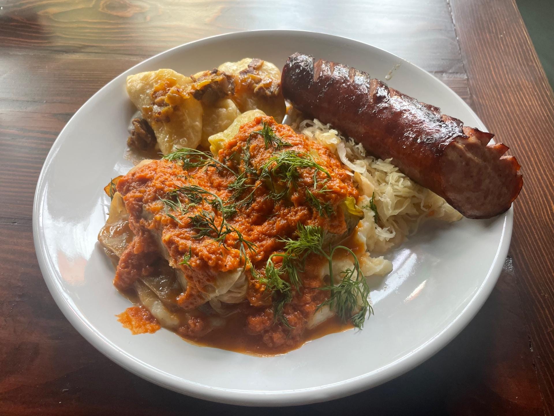 A plate of Eastern European cuisine, perfect for mastering a cozy weekend in Greene County, features a grilled sausage, stuffed cabbage rolls topped with tomato sauce and fresh dill, served with a side of sauerkraut on a rustic wooden table.