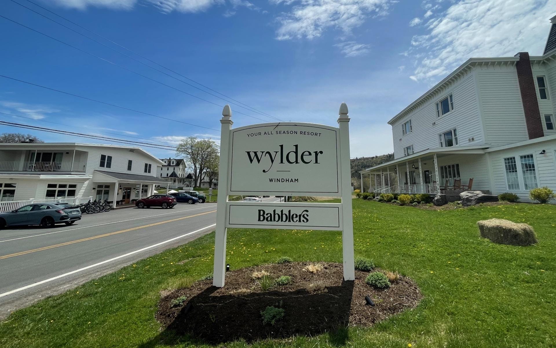 A roadside sign reads Wylder Windham and Babblers, perfectly capturing the essence of a weekend escape. It stands on a grassy lawn with a few small plants, in front of a white building along the road in Greene County, under a partly cloudy sky.