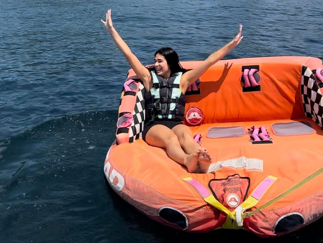 A woman in a life vest sits on an inflatable raft with her arms raised, smiling widely. The raft, floating on the calm waters of Lake George, boasts a bright orange and pink checkered design.