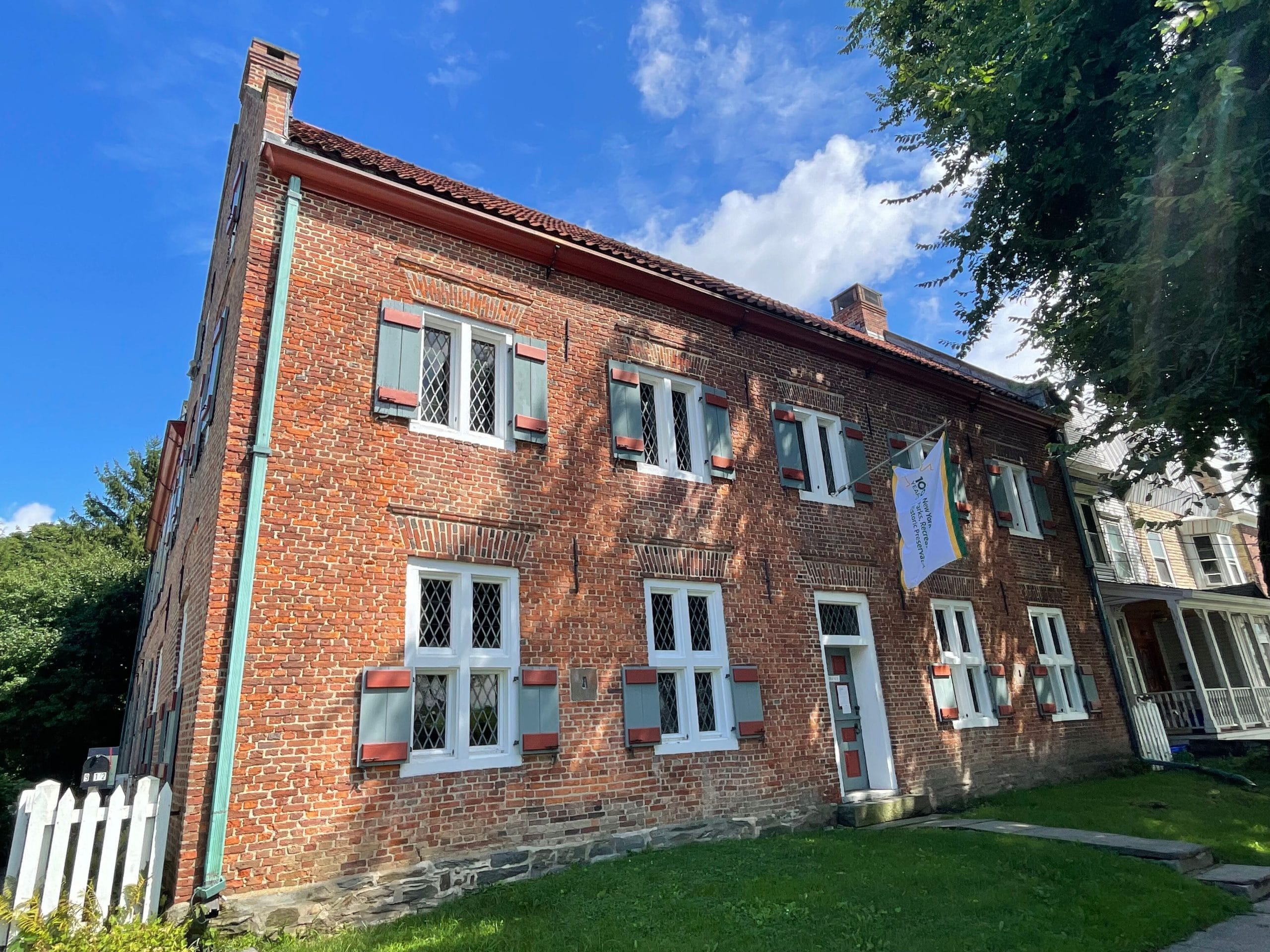 A historic brick building with green and red shutters stands under a partly cloudy sky, exuding timeless beauty. A white banner hangs near the entrance, and a white picket fence borders a lush green lawn in the foreground—a picturesque scene reflective of New York State Parks.