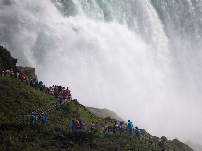 A large group of people in raincoats stand on a wooden walkway by a powerful waterfall within New York State Parks. The mist creates a dramatic backdrop, highlighting the beauty of natures wonder, while lush greenery surrounds the path.