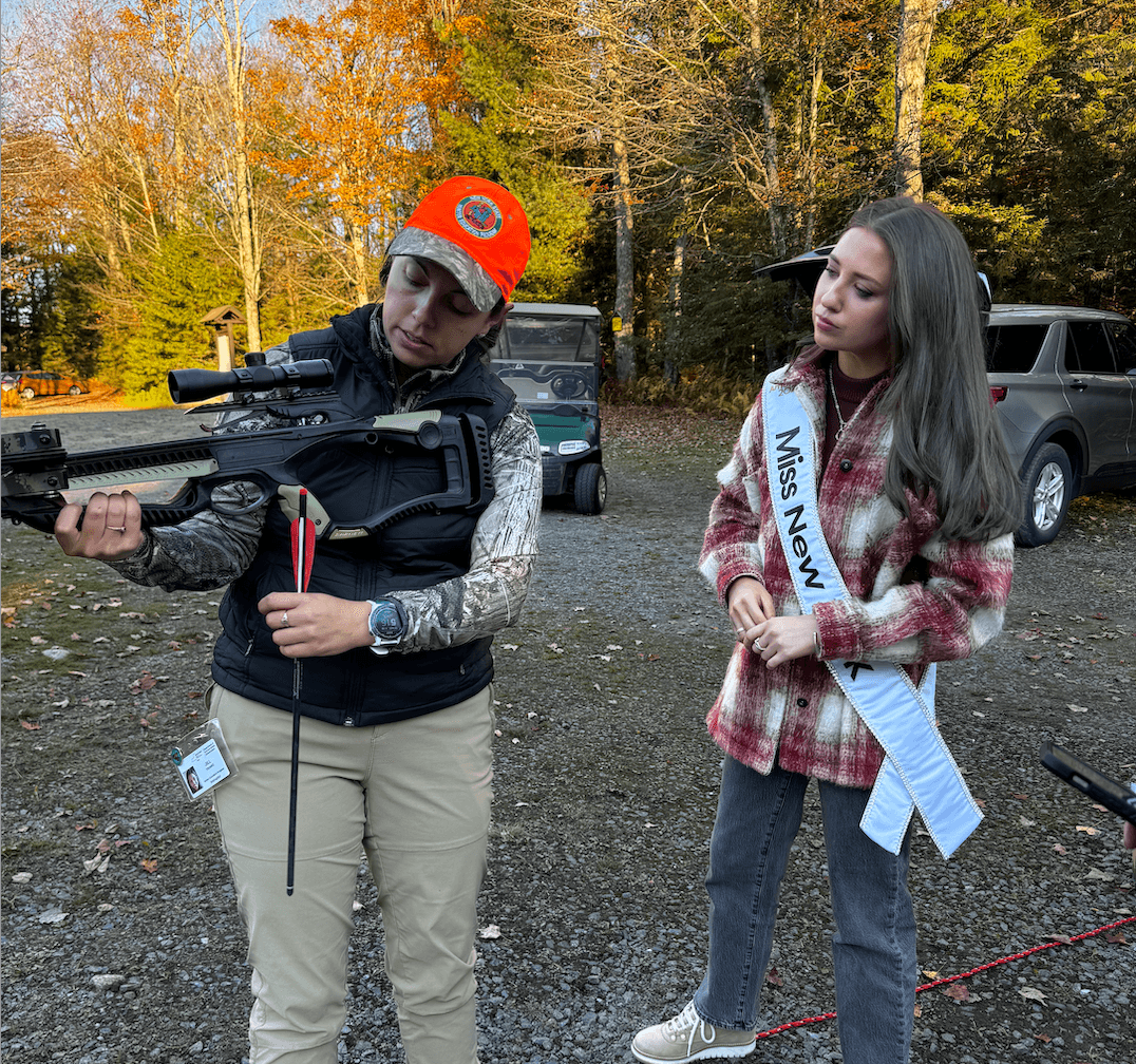 Two women are outdoors in the Adirondacks by a wooded area. One is holding and examining a crossbow with arrows. The other, wearing a Miss Nevada sash, looks on attentively. The scene is surrounded by trees adorned with vibrant autumn leaves.