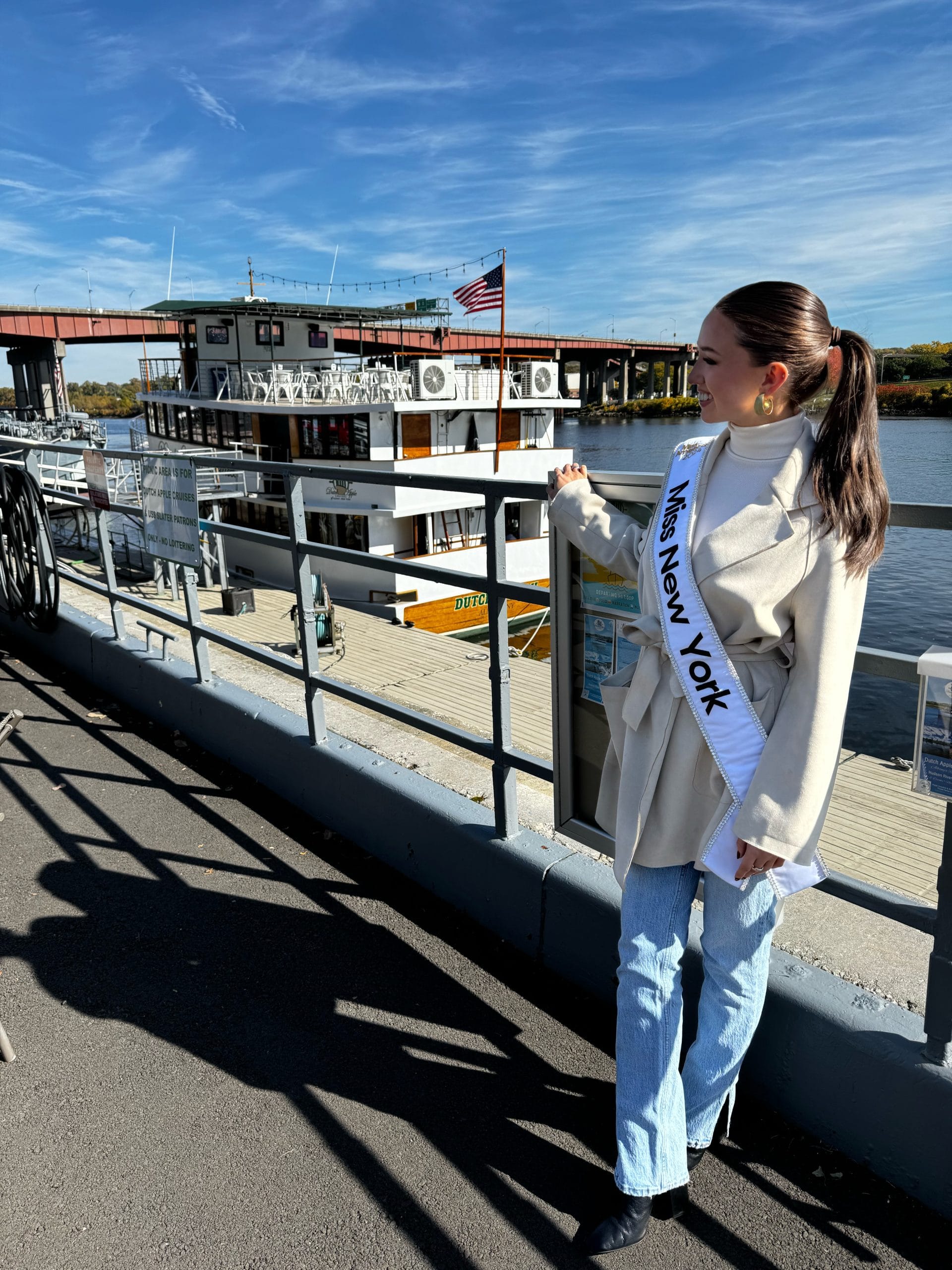 A woman wearing a Miss New York sash stands by a railing in Albany, admiring a riverboat adorned with an American flag. She is dressed in a beige coat and jeans, under the clear blue sky.
