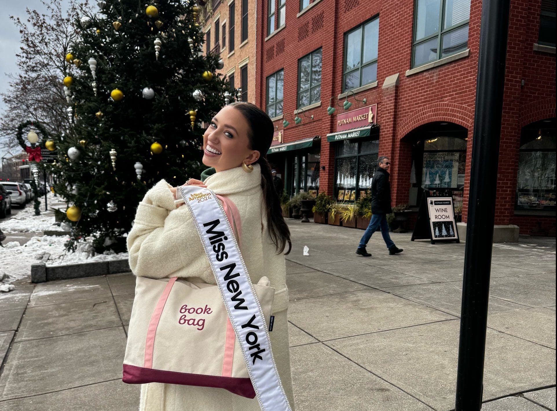 Abigail Quammen, Miss New York, stands on a snowy Saratoga sidewalk, smiling and holding a tote bag labeled book bag. She is proudly wearing her Miss New York sash draped over her shoulder. A decorated Christmas tree and charming brick buildings complete the festive backdrop.