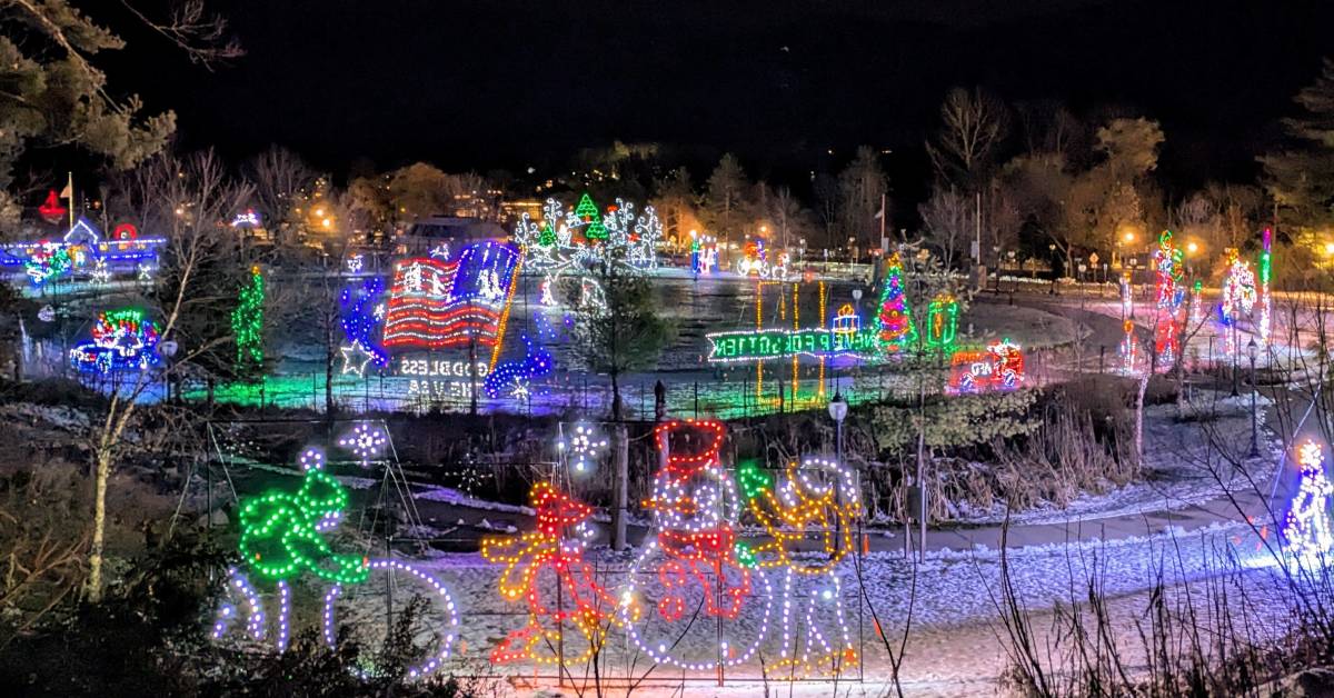A vibrant holiday light display, part of the Lights at the Lake event, illuminates a snowy park at night. Various festive shapes, including snowmen, trees, and a waving American flag, are formed with colorful lights. The scene creates a cheerful and festive atmosphere.