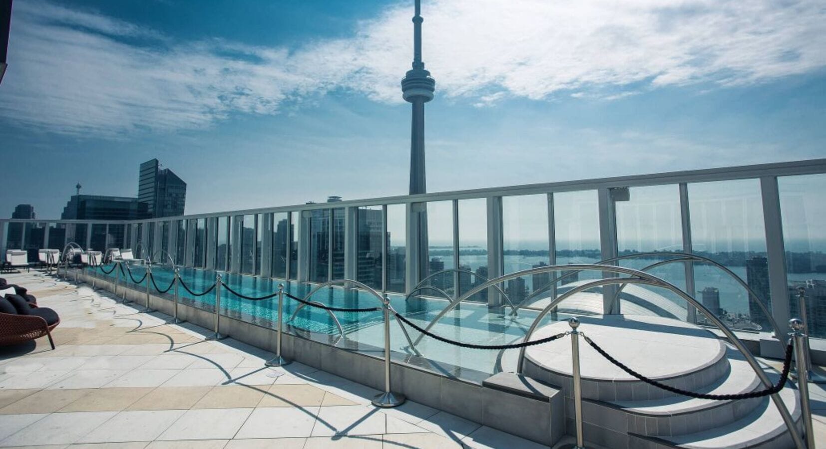 Rooftop view at Bisha Hotel featuring an outdoor pool with glass railings overlooking a cityscape. The CN Tower is prominently visible against a partly cloudy sky. Lounge chairs are placed by the pool, and modern buildings are in the background.