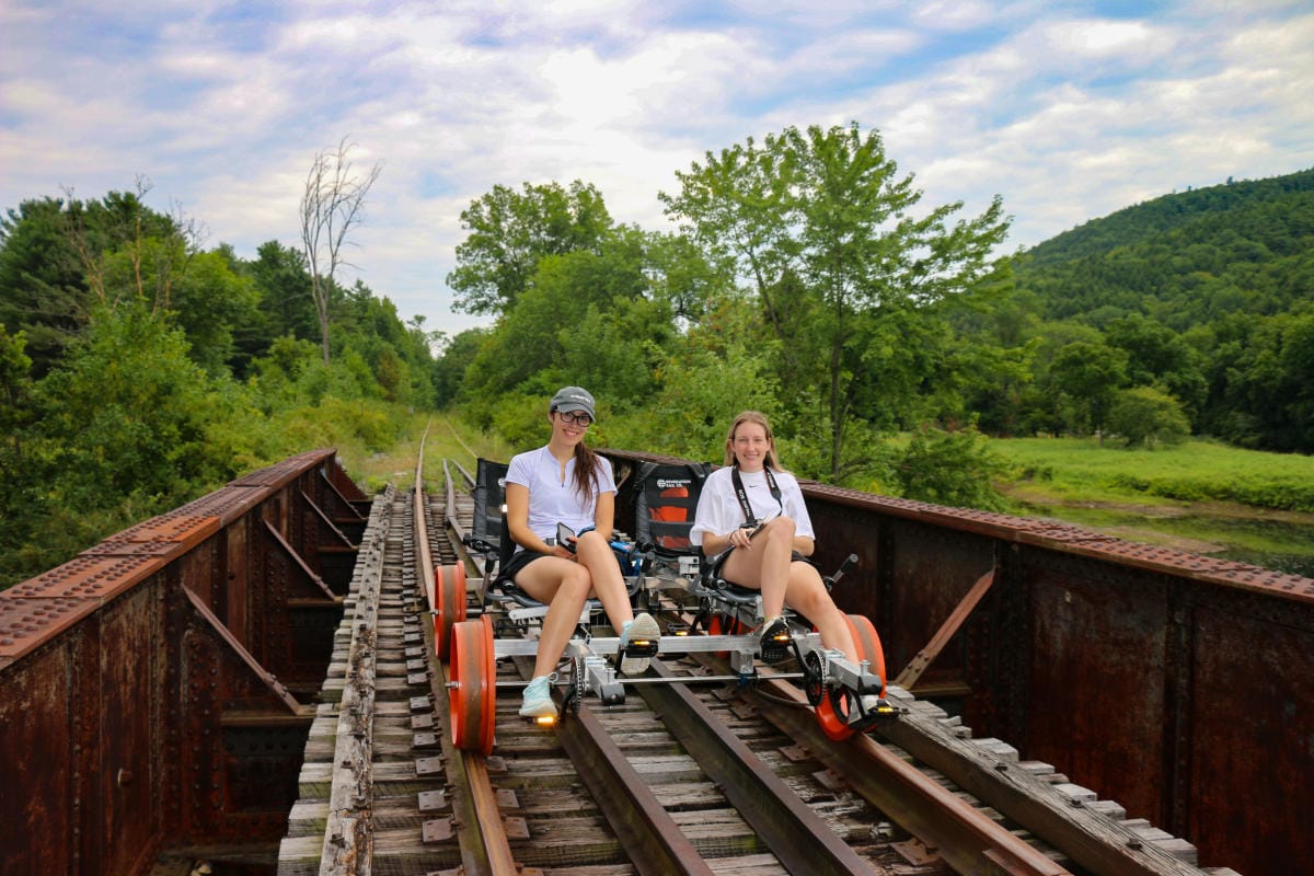 Two people are seated on a railbike, riding along a rusty railway track near Reidlbauers Resort. Lush greenery and hills surround them under a partly cloudy sky. Both are smiling and dressed casually.
