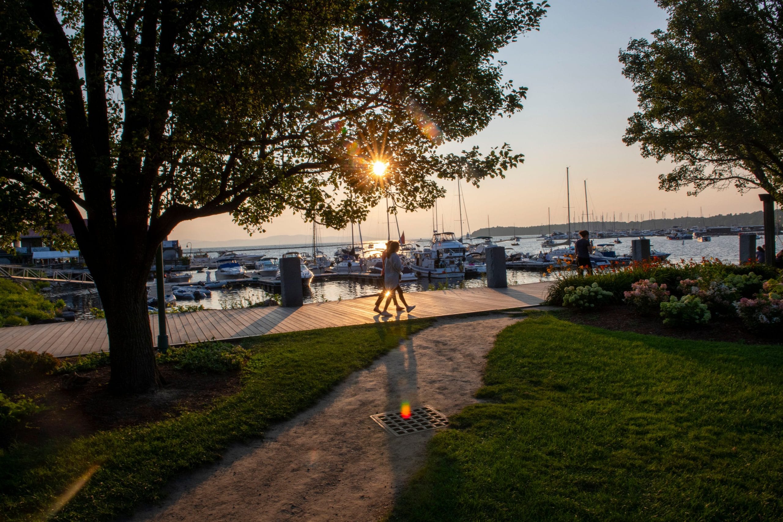 Two people walk along a sunlit path by a marina at sunset, enjoying a car-free trip. Sailboats rest in the water as trees frame the scene, with sunlight streaming through branches and casting long shadows on the boardwalk and grass.