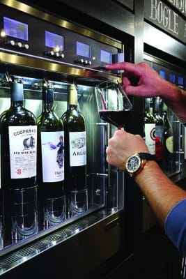 A person dispenses red wine from a wine dispensing machine into a glass at The Shaker & Vine in Schenectady holding it under the nozzle. Four wine bottles with different labels are visible inside the machine.