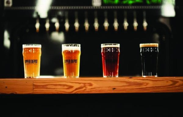 Four pint glasses at Upper Depot in Hudson New York filled with different kinds of Hudson Valley beer—ranging from pale to dark—are lined up on a wooden bar, with beer taps blurred in the background.