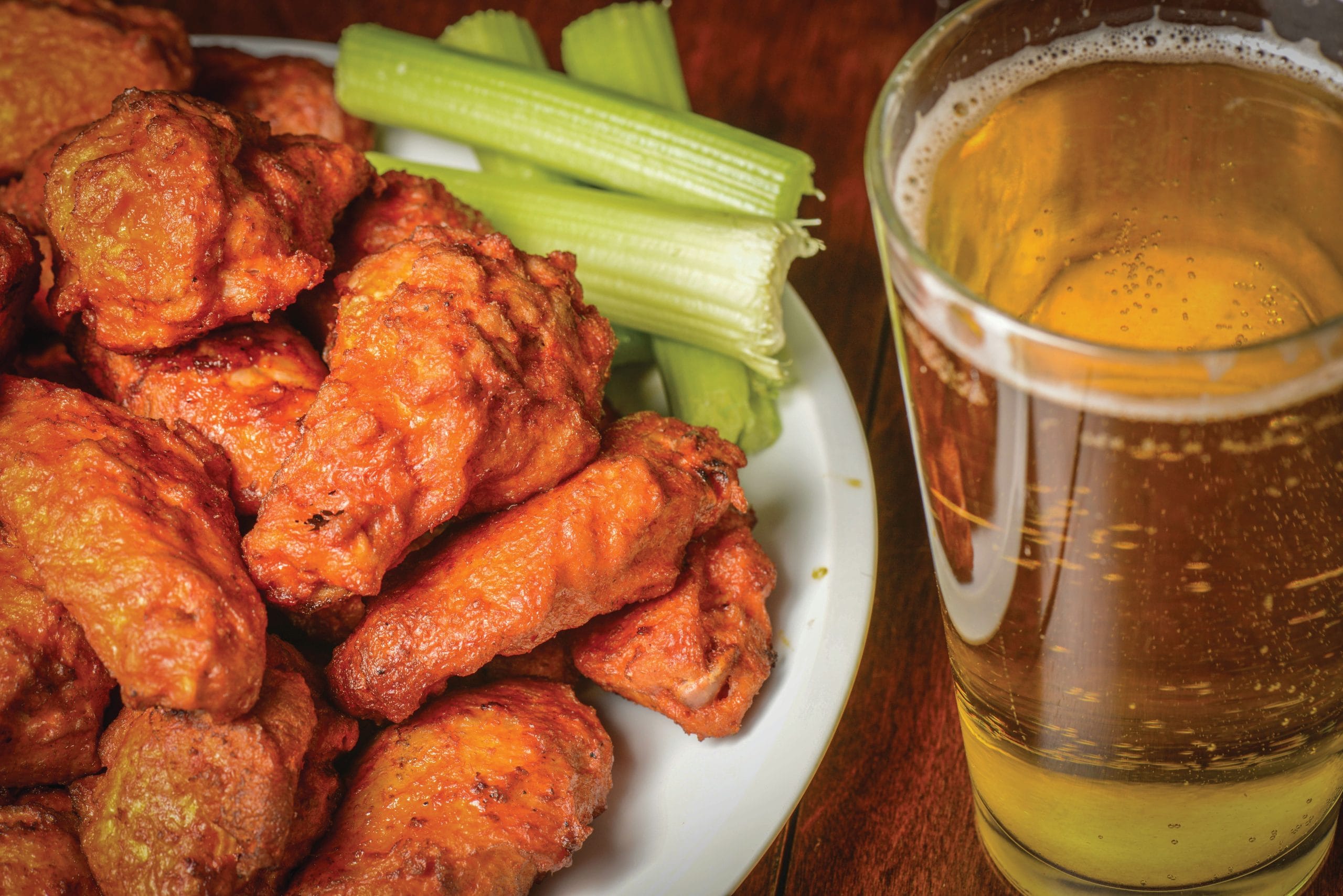 A plate of saucy buffalo chicken wings with celery sticks and a glass of beer on a wooden table—perfect for sampling classic New York regional dishes on your Empire State eats adventure.