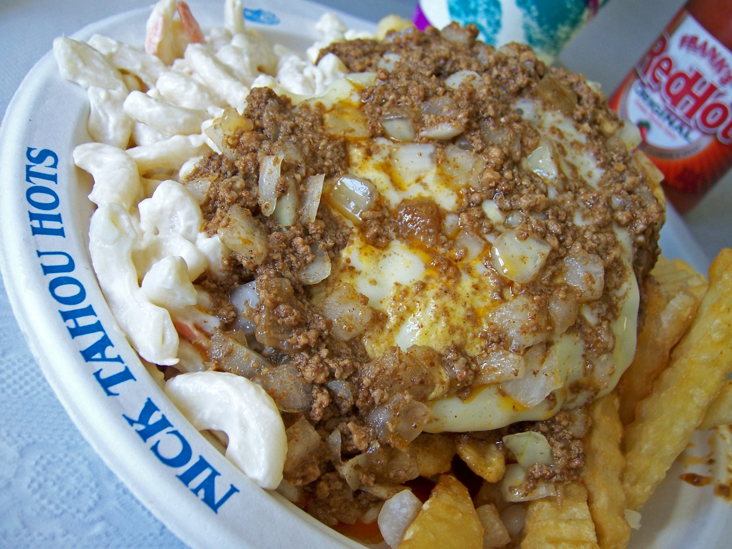 A plate of food featuring macaroni salad, crinkle-cut fries, and a hamburger patty topped with chopped onions, seasoned ground beef, and sauce—a classic among New York regional dishes—served on a Nick Tahou Hots branded dish.