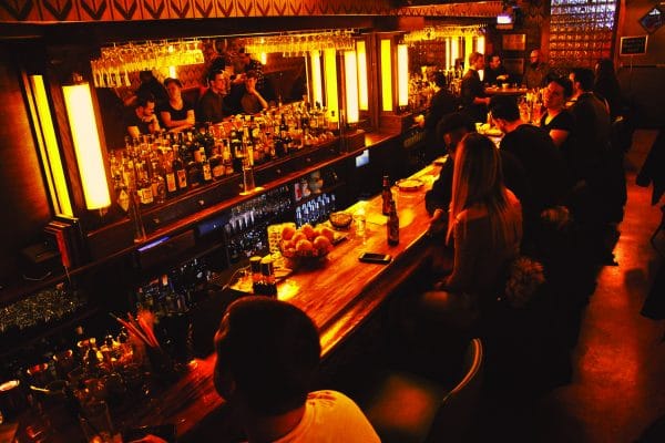 A dimly lit New York City bar with people sitting along a wooden counter, illuminated by warm yellow lights. Bottles and glasses line the shelves behind the bar as patrons enjoy conversation and drinks.